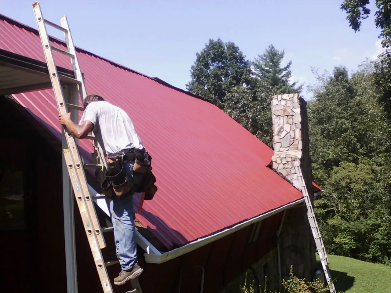 Roofer on a ladder beside a red metal roof and stone chimney — Soffit & Fascia Repair by Roofing Brothers in Crooks